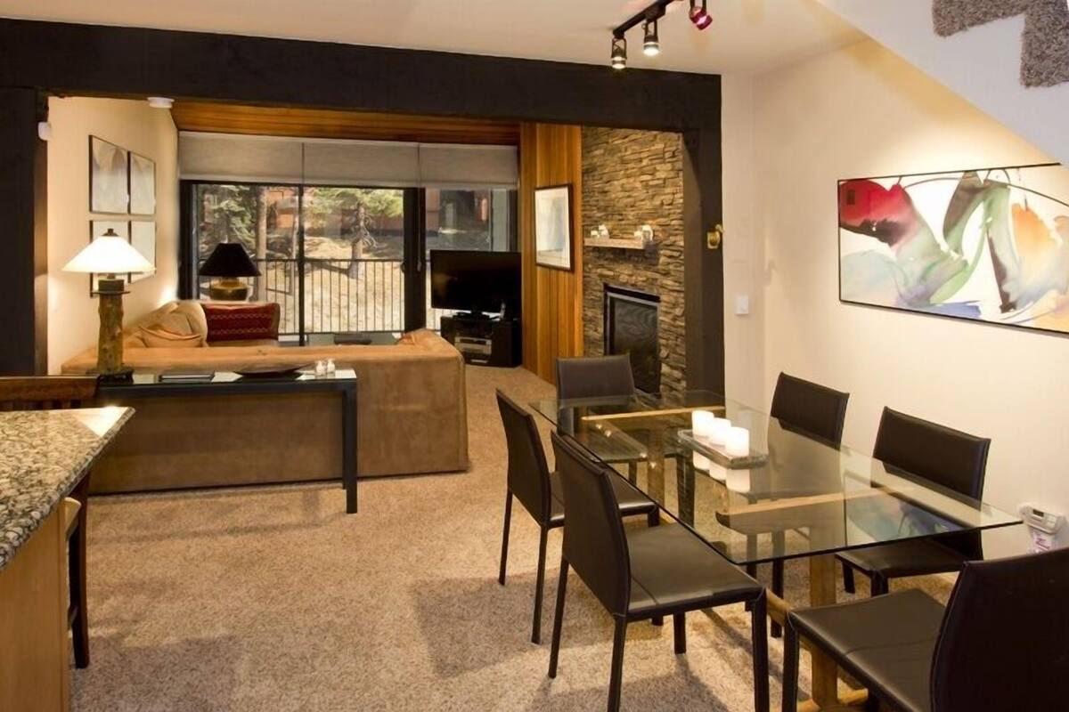A modern living room with a glass dining table, comfortable seating, a fireplace, and a large window offering a view of the outdoors.
