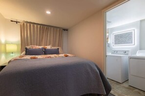 A bedroom with a bed, a nightstand, a lamp, a window with curtains, and a laundry area with washing machines.