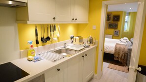 A compact kitchen with white cabinets, a built-in stove, and a sink. A toaster and kettle are on the counter. The room has yellow walls and a door leading to a bedroom with a bed and a small table.