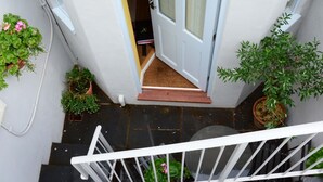 A staircase leading to a door with a small landing area. There are potted plants on both sides of the staircase and a white railing in the foreground.