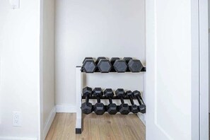 Un coin avec un mur blanc et un plancher en bois, où l’on aperçoit un support mural sur lequel sont rangés plusieurs haltères.