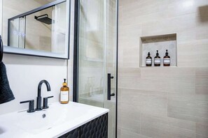 Un baño con un lavamanos blanco, cabina de ducha de cristal y tres botellas en un estante.