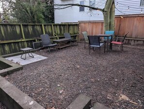 Un patio trasero con piso de concreto, cerca de madera y mobiliario exterior, incluyendo sillas y una mesa.