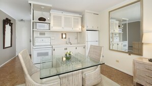 A modern kitchen with white cabinetry, a glass-top dining table, wicker chairs, and a large mirror. - Lovely Studio in the Heart of Waikiki – A/C, Wi-Fi, FREE Parking! Sleeps 2 (Honolulu)