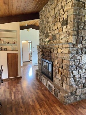 A stone fireplace with a TV, wooden floors, and a kitchen area with shelves and cabinets.