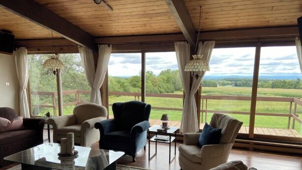 Un salon spacieux doté d’un plafond en bois, de grandes fenêtres offrant une vue sur un paysage verdoyant, et de divers sièges, notamment un canapé, des fauteuils et une table basse.