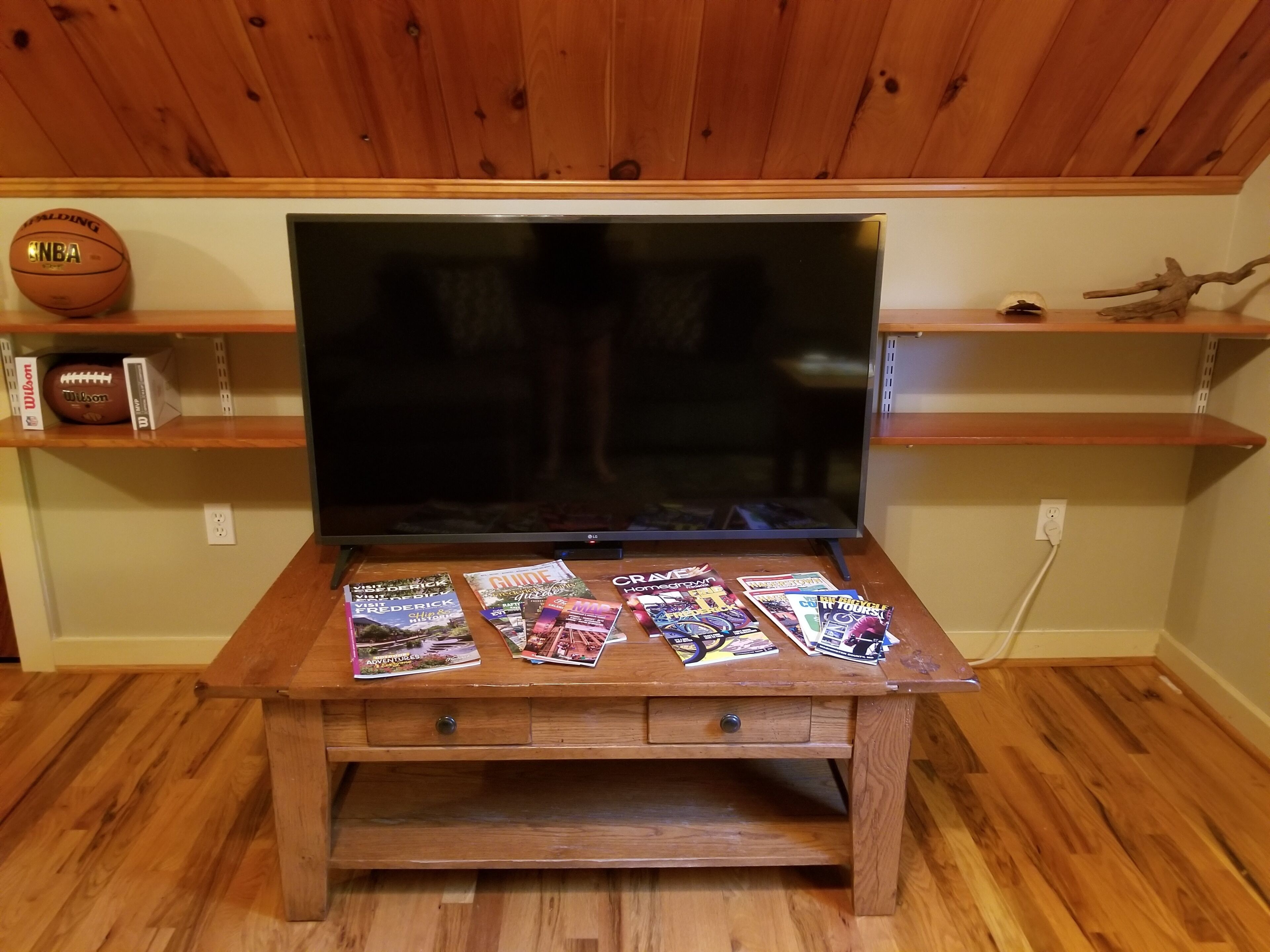 Un mueble de entretenimiento de madera con un televisor de pantalla plana, un estante con una pelota de baloncesto y un balón de fútbol, y una mesa con revistas de viajes.