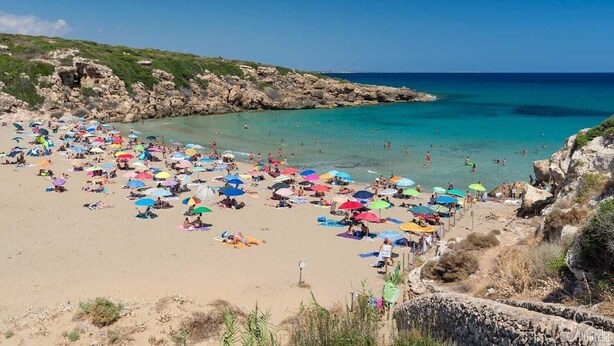 Ein überfüllter Strand mit vielen bunten Sonnenschirmen und Menschen, die das Wasser genießen.