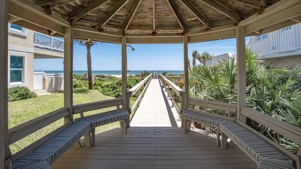 A wooden gazebo with a covered walkway leading to the beach, benches along the sides, and a view of the ocean.