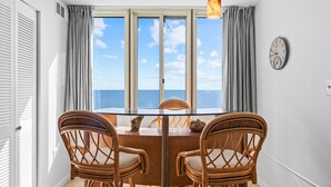 Zona pranzo con tavolo e quattro sedie in vimini, lampadario, orologio a parete e vista sul mare tramite ampie finestre.