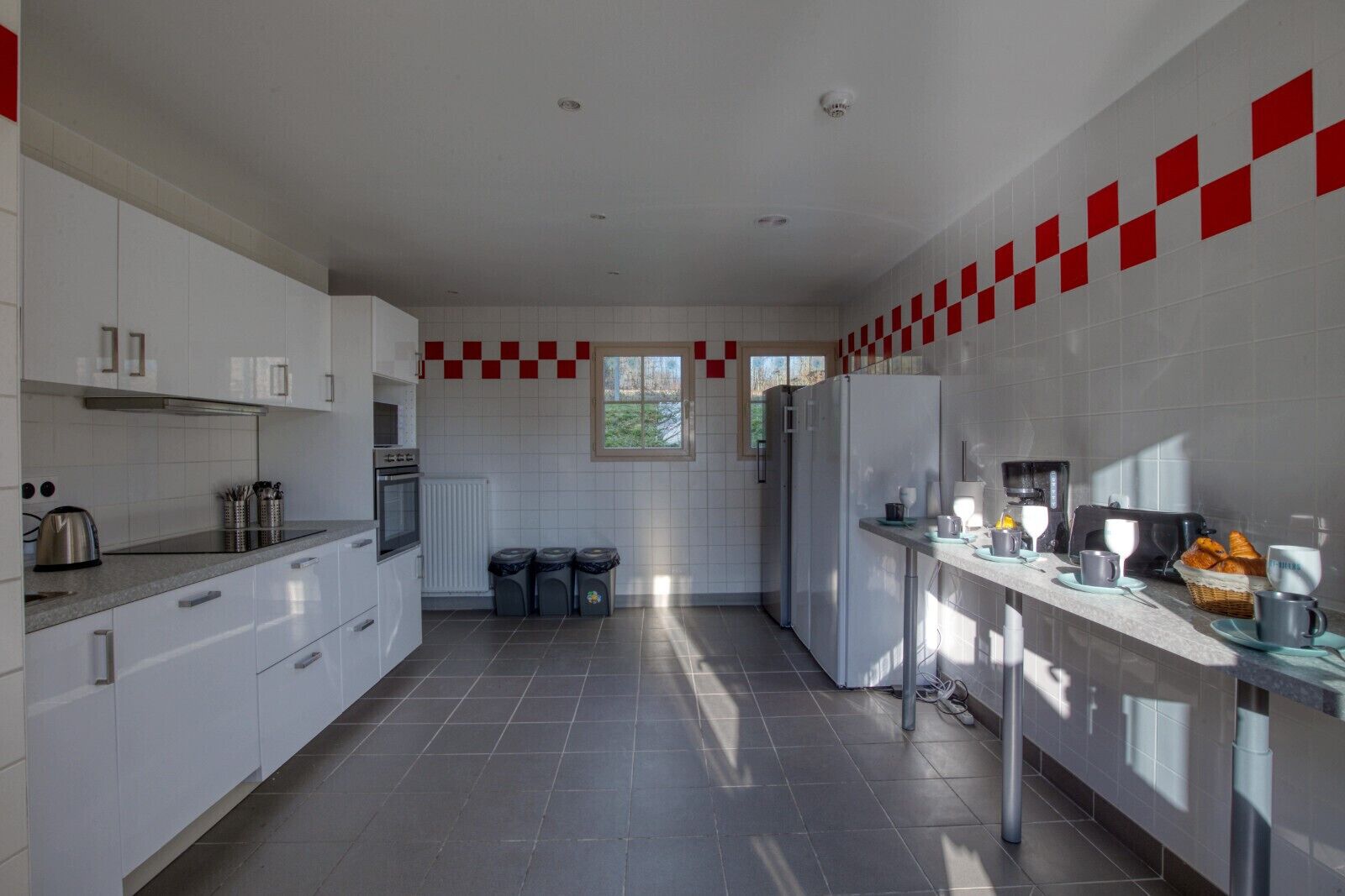 A modern kitchen with white cabinets, a built-in oven, a refrigerator, and a coffee machine. There are three trash bins and a window providing natural light.