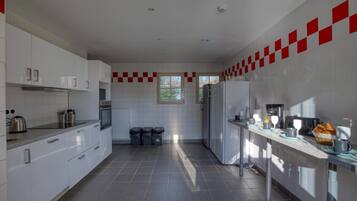 A modern kitchen with white cabinets, a built-in oven, a refrigerator, and a coffee machine. There are three trash bins and a window providing natural light.