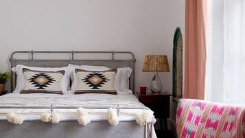 A bedroom with a metal bed frame, two pillows with geometric patterns, a woven lamp, a chair with a colorful fabric, and a hanging light fixture.