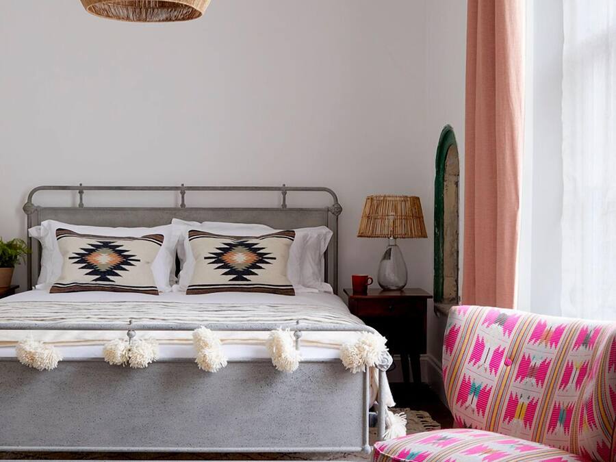 A bedroom with a metal bed frame, two pillows with geometric patterns, a woven lamp, a chair with a colorful fabric, and a hanging light fixture.