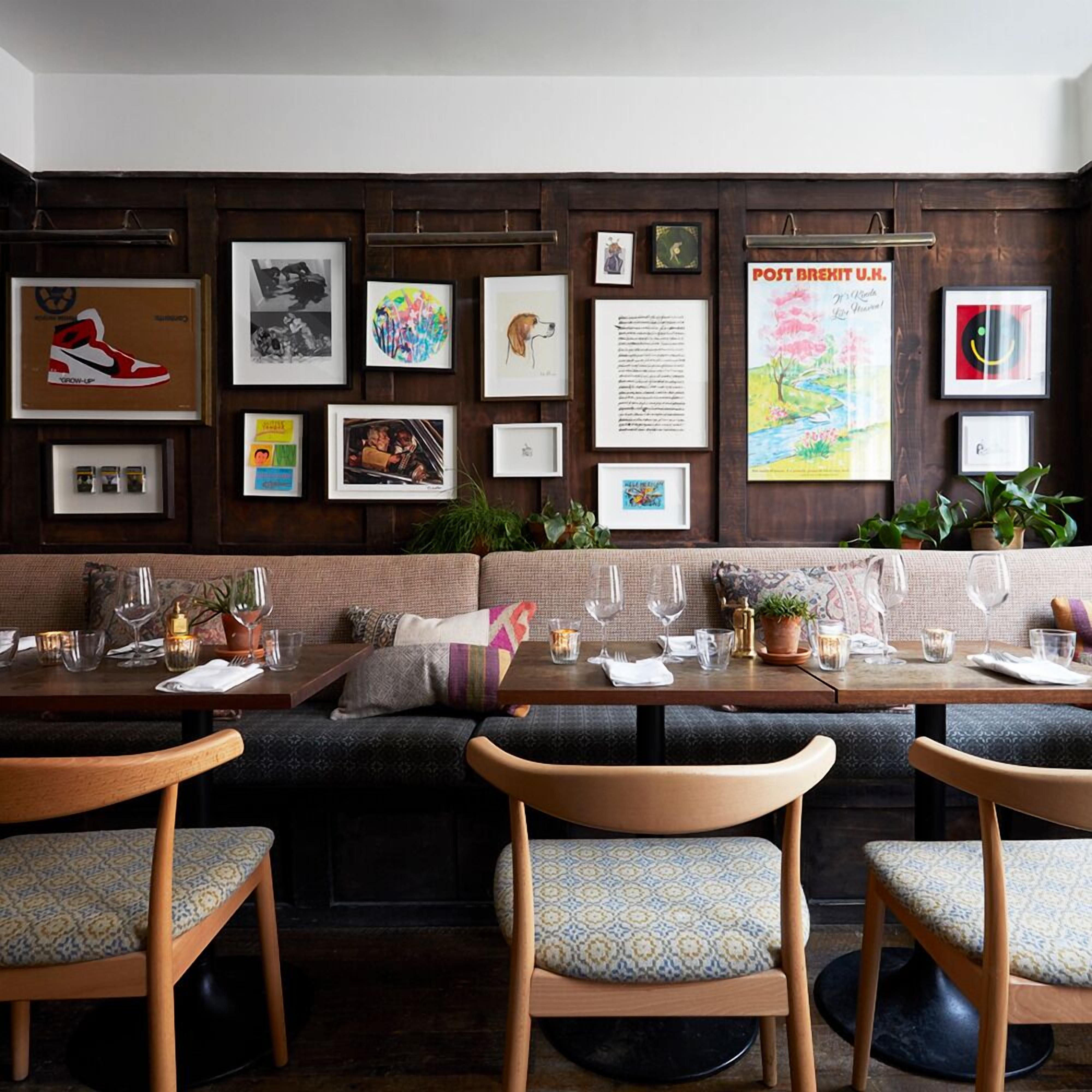 A restaurant with a long wooden wall adorned with various framed artworks, a sofa, and a dining area with wooden chairs and tables set for two.