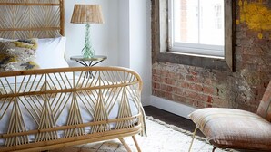 A room with a rattan bed, a small round table with a lamp, a chair, and a large window with a view of a brick building. - Artist Residence Bristol (Bristol)
