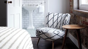 A room with a white door, a mirror above a white sink, a patterned chair, a small wooden table, and a window with a view of a brick building. - Artist Residence Bristol (Bristol)