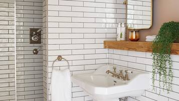 Un baño con paredes de ladrillo blanco, un lavabo blanco, un espejo, una ducha y una planta colgante.