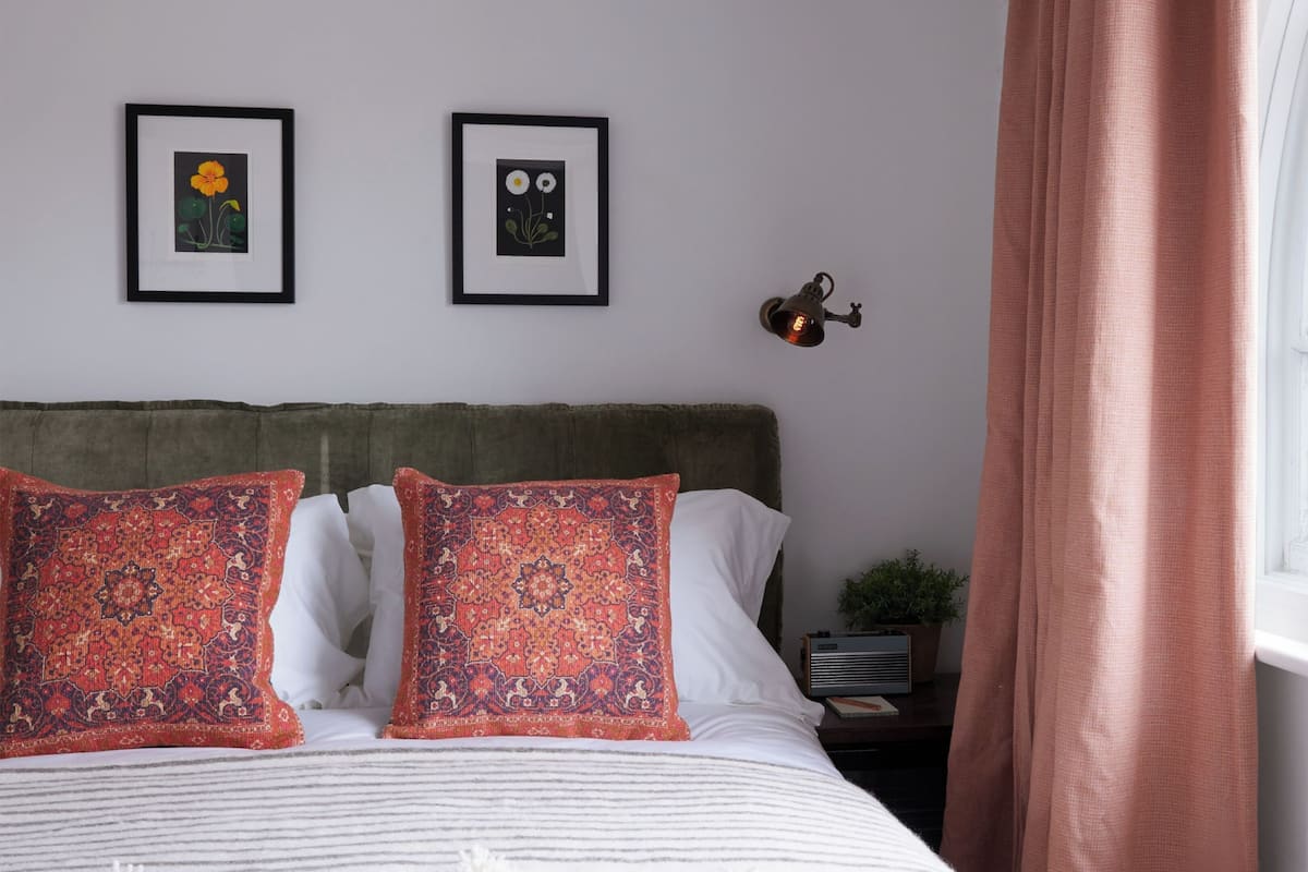 Ett sovrum med en säng, två inramade konstverk med blommotiv, ett sängbord, en lampa och ett fönster med gardiner.