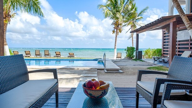 A poolside area with lounge chairs, a table with a bowl of fruit, and a view of the ocean. - Beautiful beach level condo, perfect for a family, pool access, WiFi, AC! (Akumal)