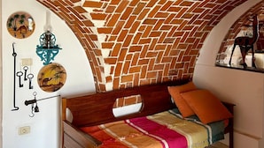 A room with a brick-patterned ceiling, a bed with colorful bedding, and wall decorations including a circular picture and a wall-mounted light fixture. - Dream room (Hergla)