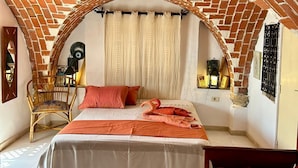 A room with a brick arch ceiling, a bed with a white and orange bedspread, a wooden chair, and a small table with a lamp. - Dream room (Hergla)