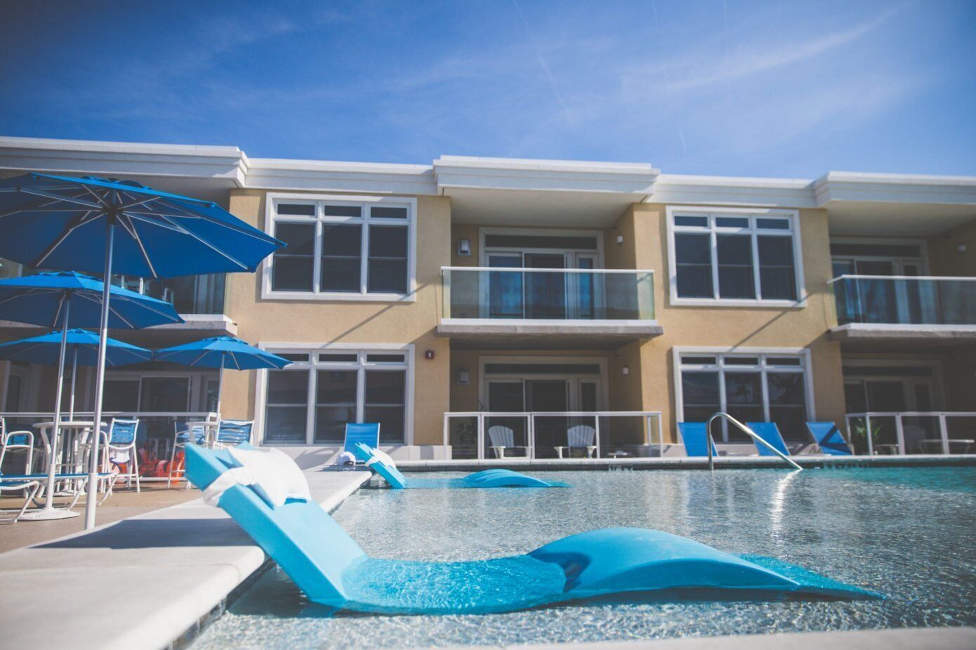 A swimming pool area with lounge chairs and umbrellas, a building with balconies, and a clear blue sky.