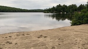 Une plage de sable où l’on peut voir des empreintes de pas menant à un lac paisible, bordé d’arbres et entouré de quelques maisons au loin.