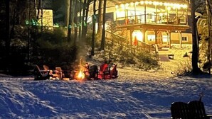 Une cabane dotée d’une grande terrasse et d’un foyer extérieur, le tout dans un paysage enneigé.