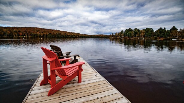 Une jetée en bois avec deux chaises Adirondack rouges et une table, d’où l’on peut admirer un lac paisible, avec des arbres et des collines en arrière-plan.