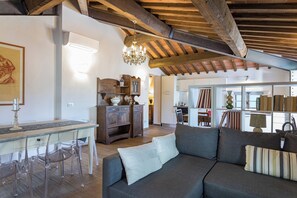 A spacious living area with a wooden ceiling, a dining table with chairs, a sofa with cushions, and a kitchen area in the background. - Antica Villa Merelli 24, Emma Villas (Poggio Fabbrelli)