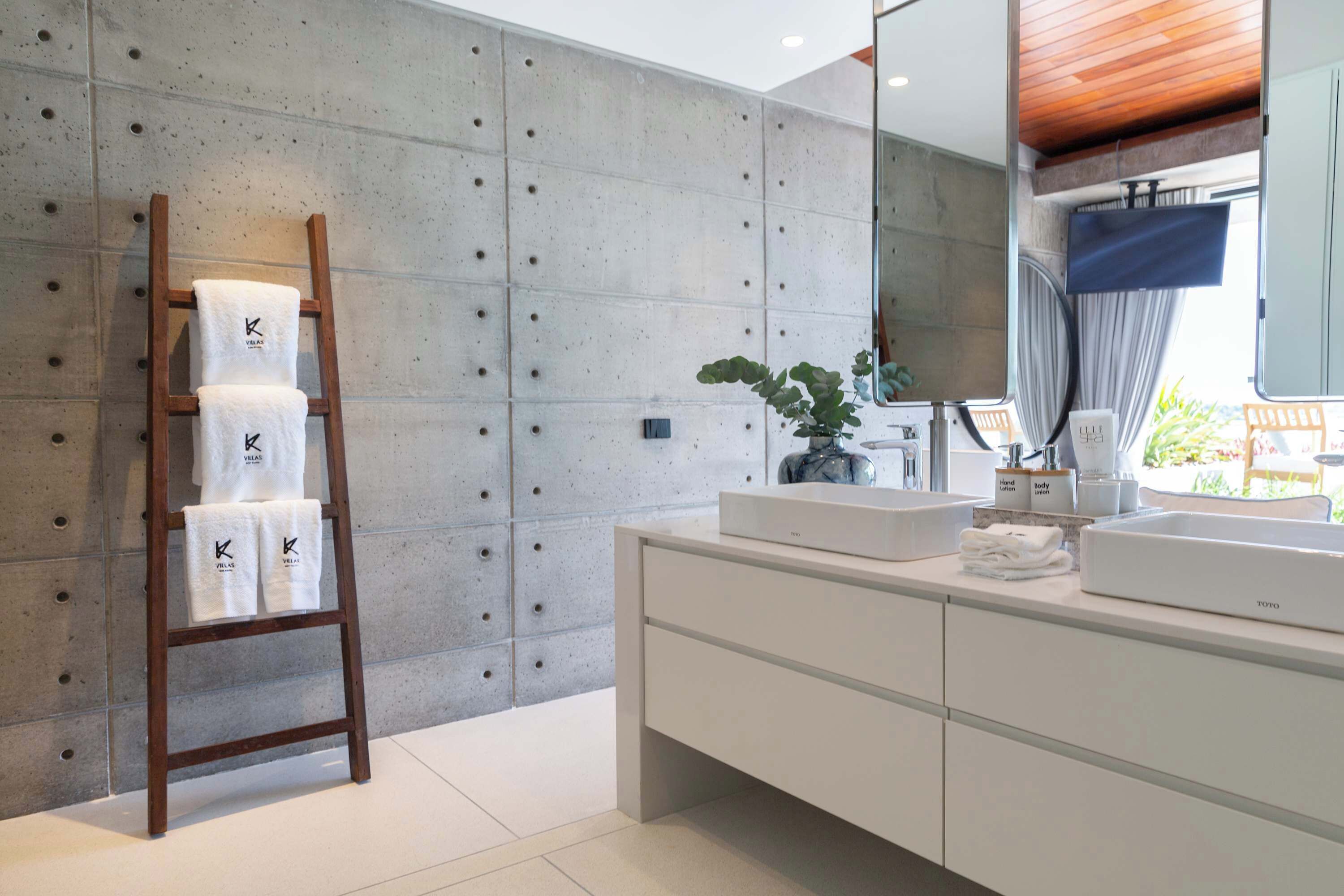 A modern bathroom with a concrete accent wall, a wooden towel rack, a large mirror, and a white vanity with a plant.