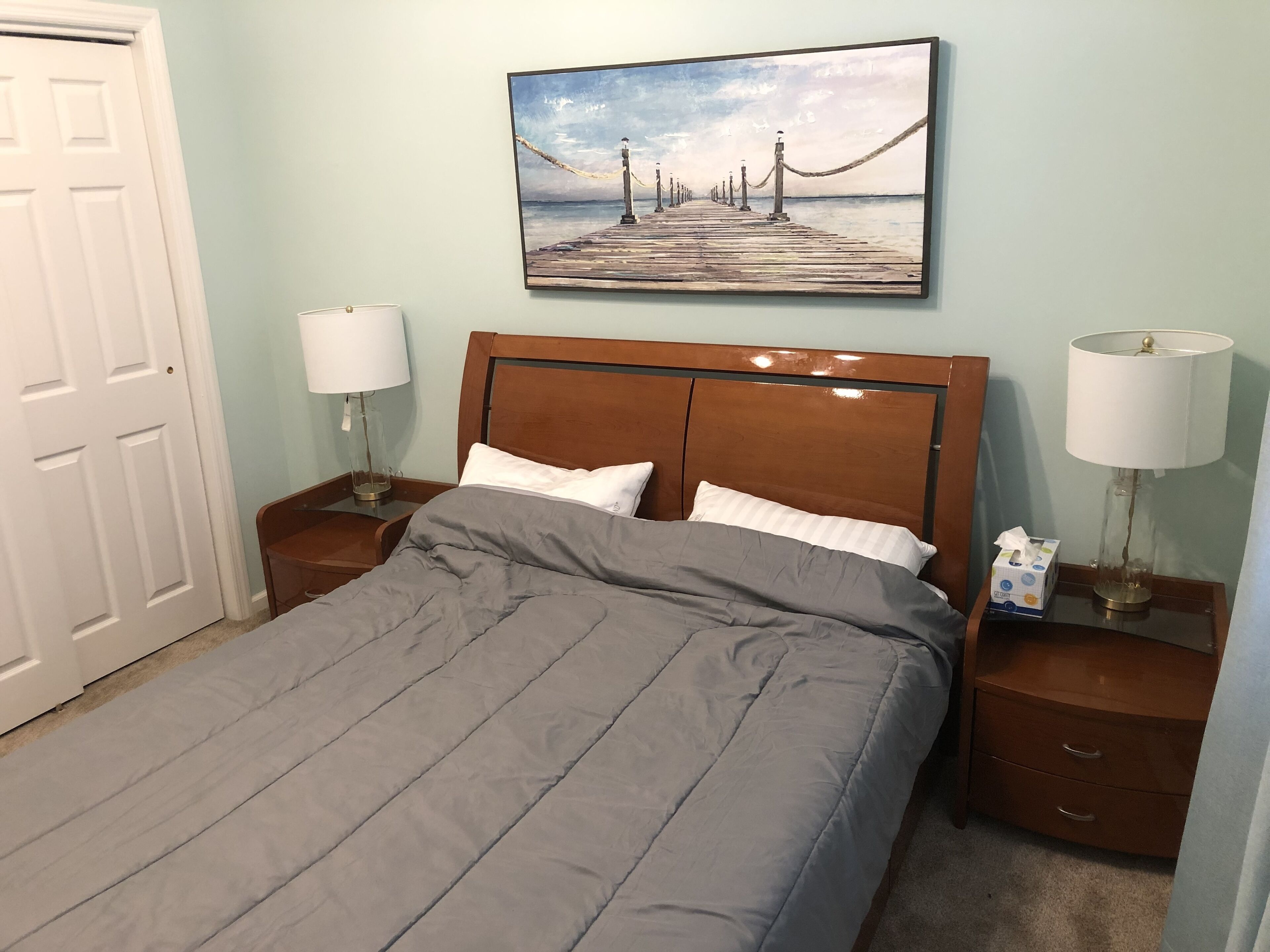 Cama bem arrumada com edredom cinza e almofadas brancas, com mesas de cabeceira de madeira, luminárias e caixa de lenços.