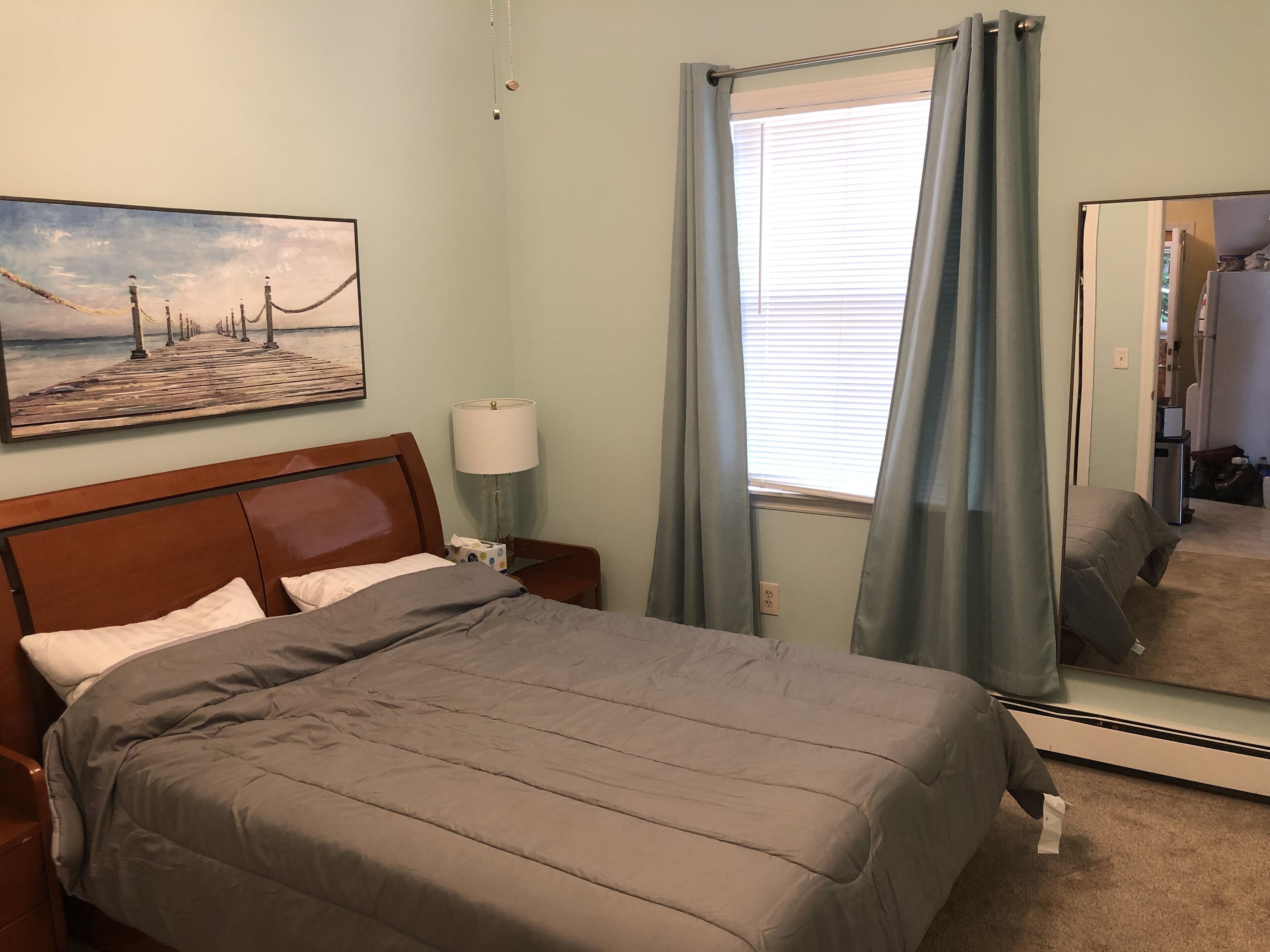 Habitación de hotel con cama, mesita de noche, lámpara, un cuadro, ventana con cortinas y espejo.