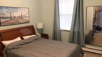 Habitación de hotel con cama, mesita de noche, lámpara, un cuadro, ventana con cortinas y espejo.