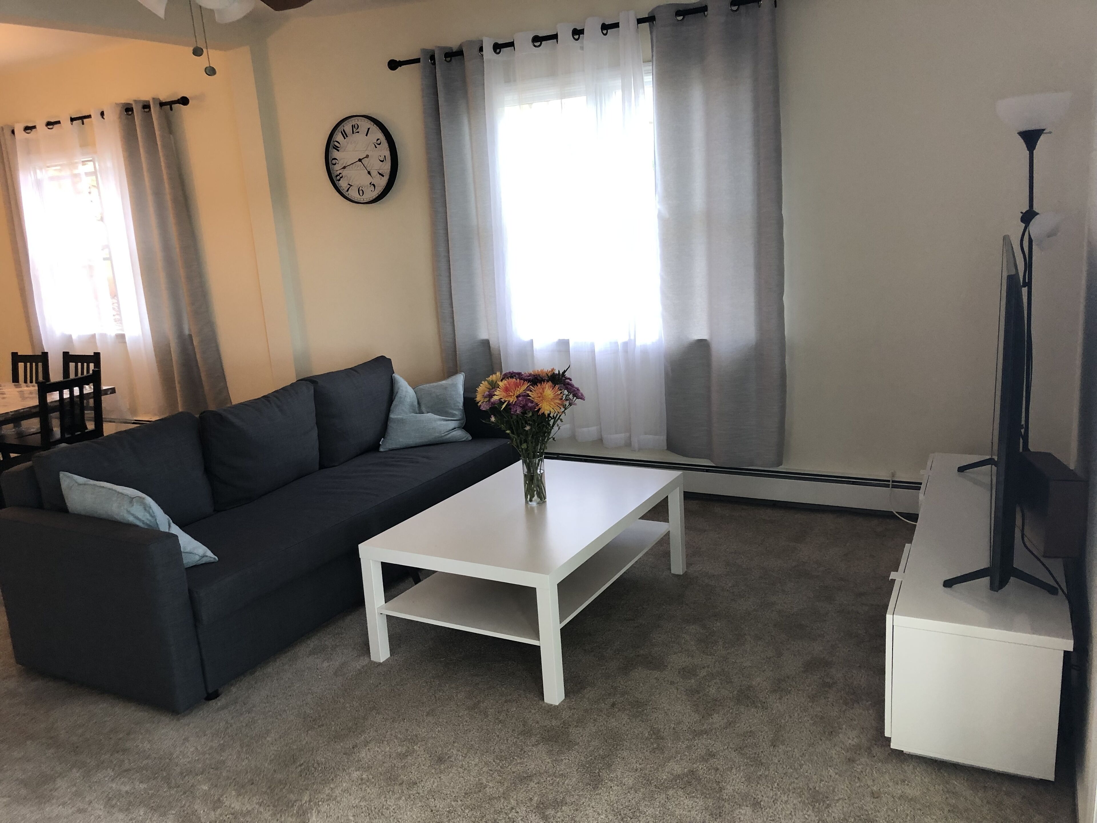 Un salon avec un canapé gris foncé, une table basse blanche, une horloge au mur et une lampe.