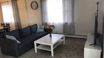 Un salon avec un canapé gris foncé, une table basse blanche, une horloge au mur et une lampe.