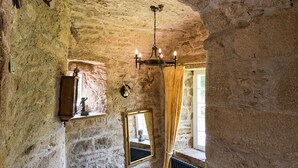 A stone-walled room with a bed, a chandelier, a mirror, and a window with curtains.