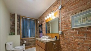 Een badkamer met een bakstenen muur, houten kastjes, een wit toilet en een wastafel met een spiegel erboven.