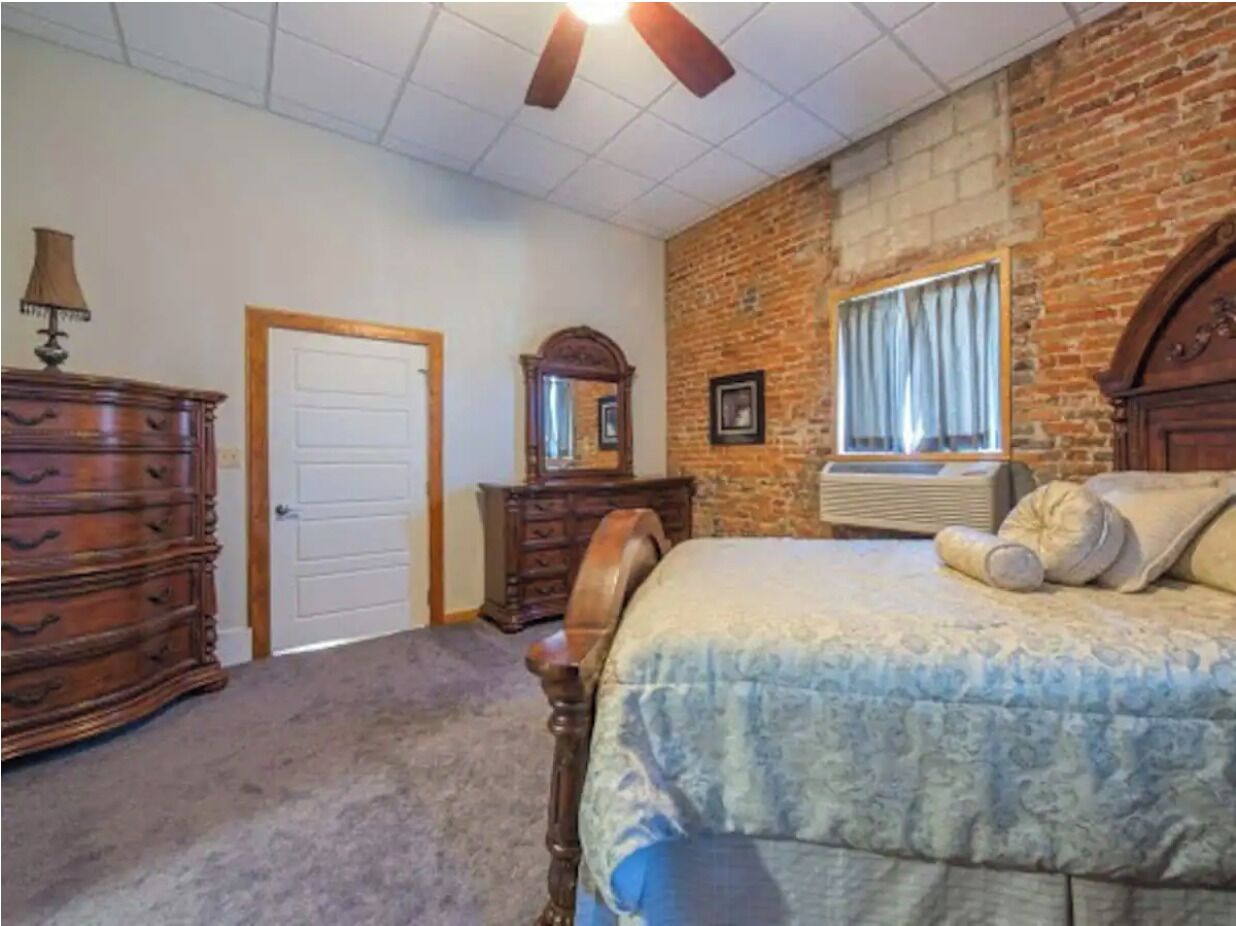 A bedroom with a brick wall, a bed with a patterned bedspread, a dresser, a mirror, and a ceiling fan.