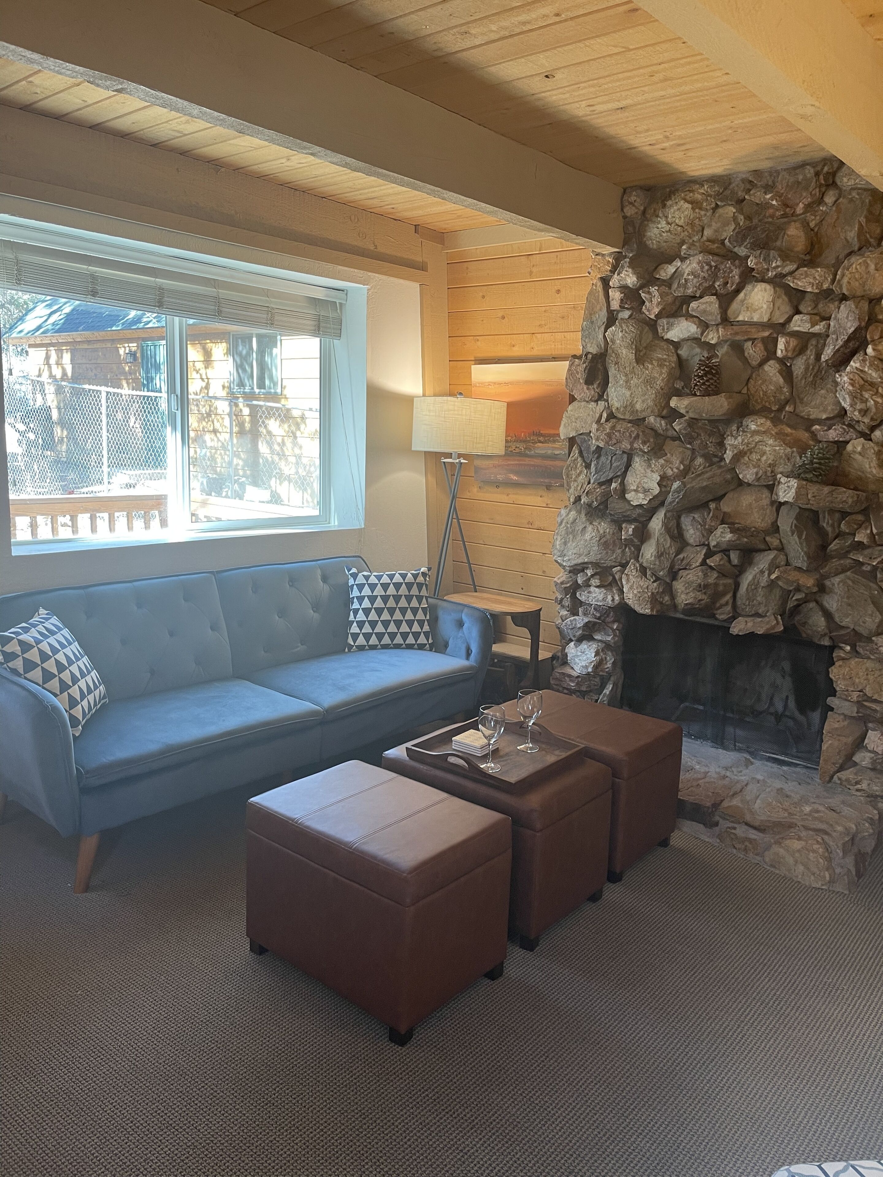 A cozy living room with a stone fireplace, a blue sofa, and two brown ottomans.