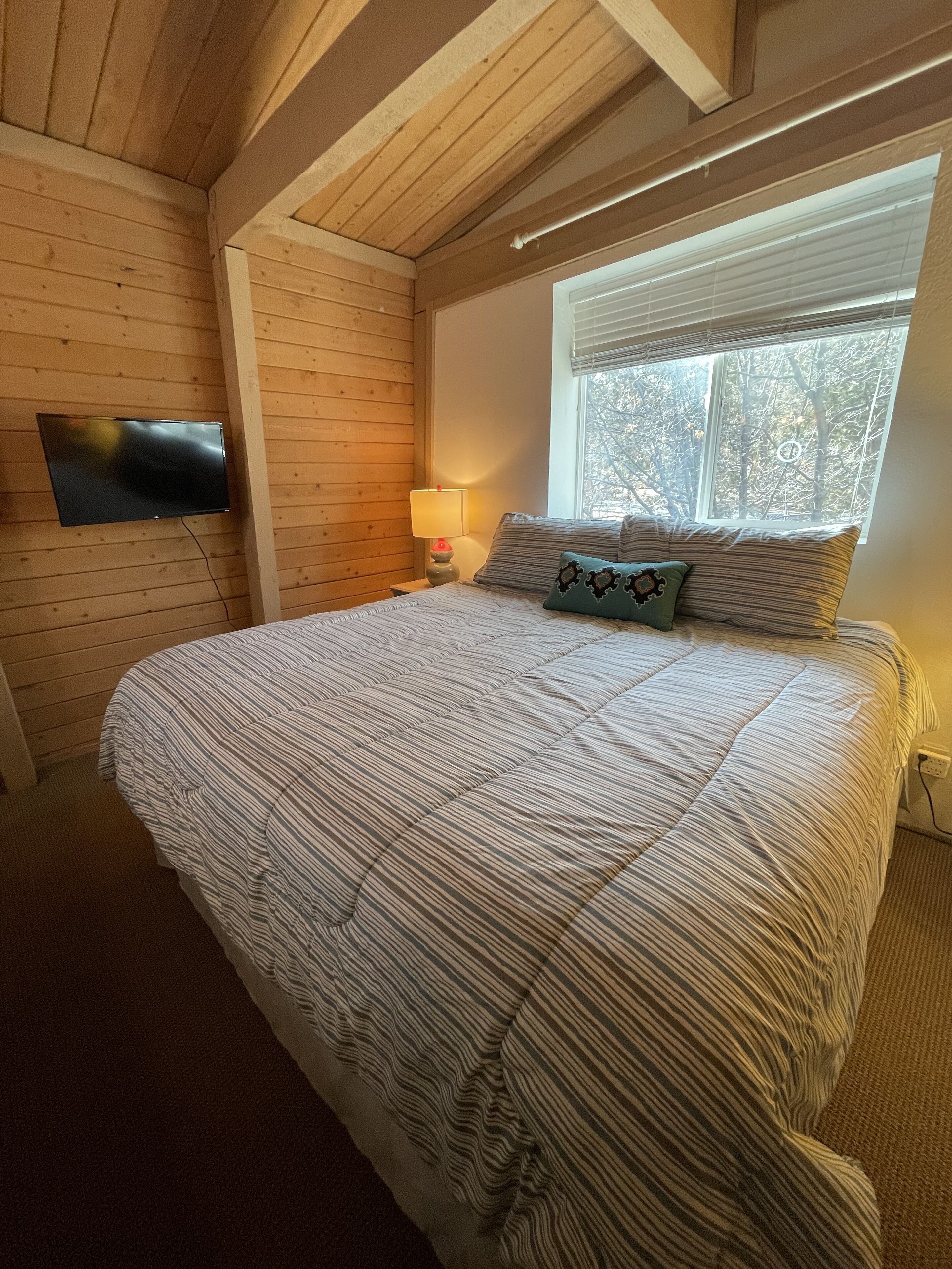 Un dormitorio con cama, televisor montado en la pared, ventana con vistas a árboles y lámpara en la mesita de noche.
