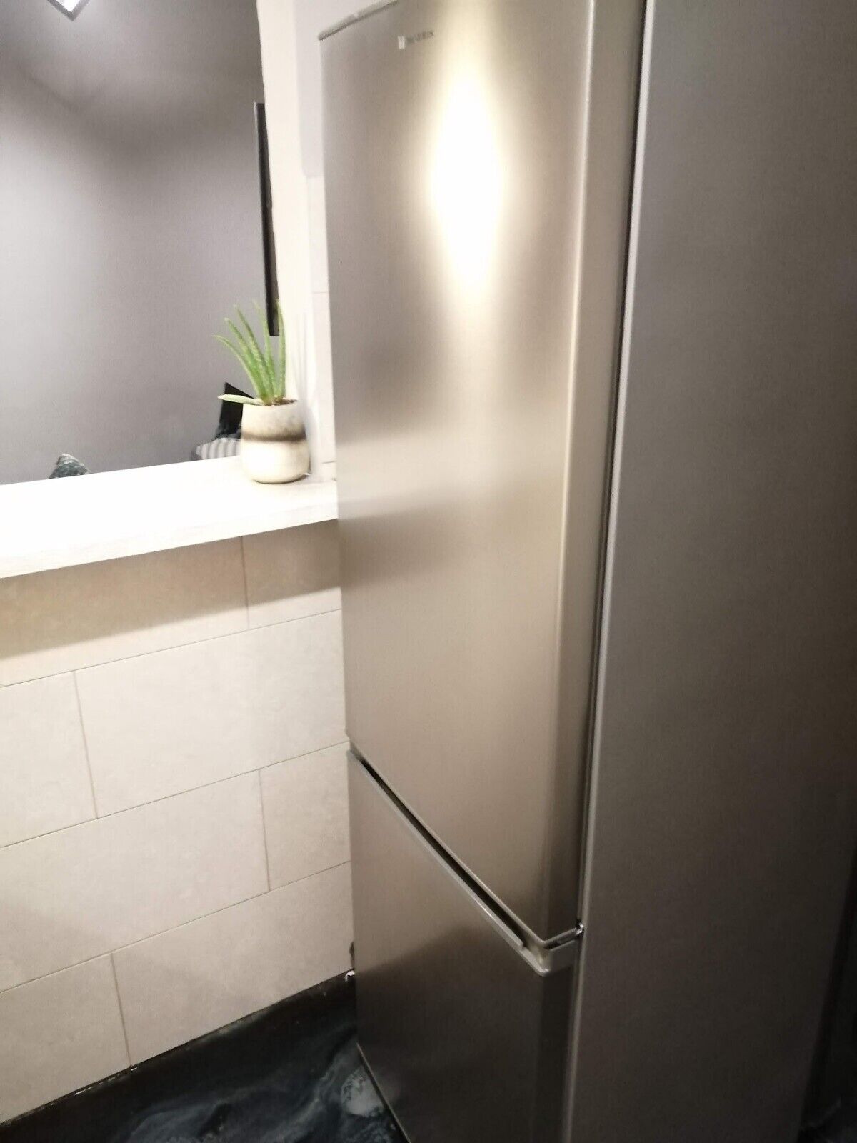 A stainless steel refrigerator in a kitchen with white tiles and a small potted plant on the counter.