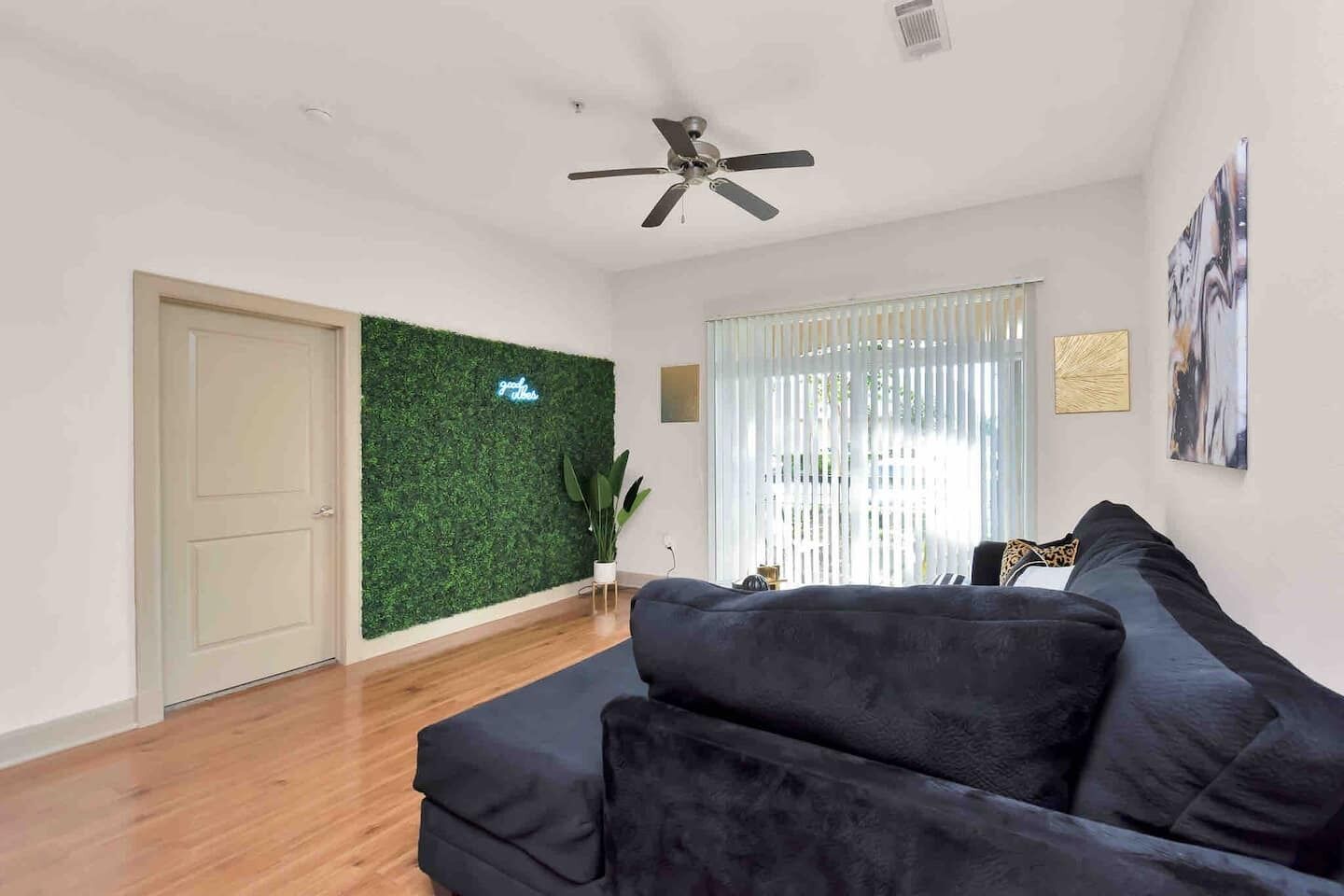 Ein modernes Wohnzimmer mit einem dunkelgrauen Sofa, einem Deckenventilator, einer grünen Wandgestaltung und einem Fenster mit transparenten Vorhängen.