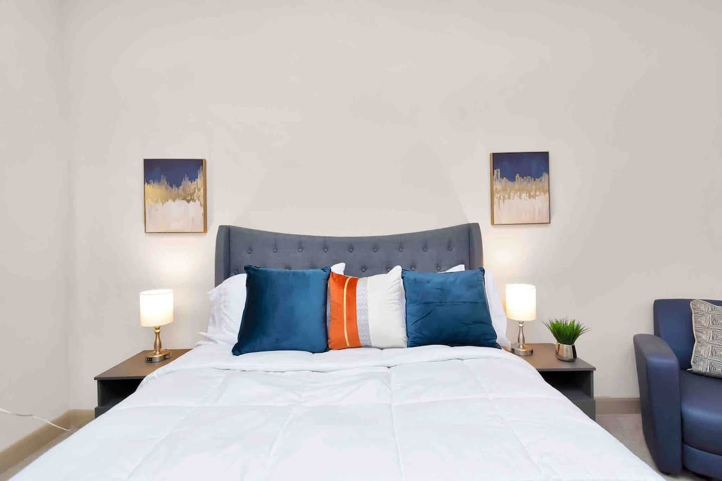 A neatly made bed with a grey upholstered headboard, white bedding, and multiple pillows in blue, orange, and white. There are two bedside tables with lamps and a small potted plant.