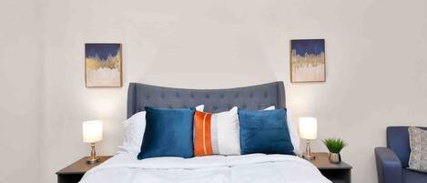 A neatly made bed with a grey upholstered headboard, white bedding, and multiple pillows in blue, orange, and white. There are two bedside tables with lamps and a small potted plant.