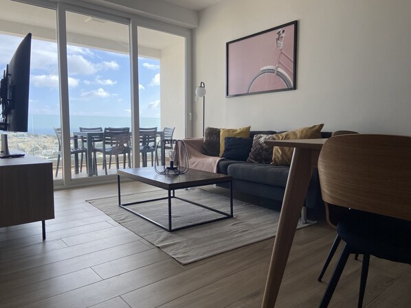 A modern living room with a sofa, coffee table, and a TV on a desk. There is a balcony with a view of the city and sky.