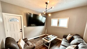 A living room with a flat-screen TV, two leather sofas, a coffee table, and a decorative light fixture. - Cozy, 2-bedroom vacation home. 1 mile to DT: Hot Tub, 86" TV, Golf & Fenced Yard (Sioux Falls)