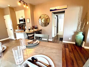 A dining area with a table set for four, a kitchen in the background, and a decorative wall piece. - Cozy, 2-bedroom vacation home. 1 mile to DT: Hot Tub, 86" TV, Golf & Fenced Yard (Sioux Falls)