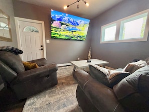 A living room with a flat-screen TV, a white door, a window, and a brown leather couch. - Cozy, 2-bedroom vacation home. 1 mile to DT: Hot Tub, 86" TV, Golf & Fenced Yard (Sioux Falls)